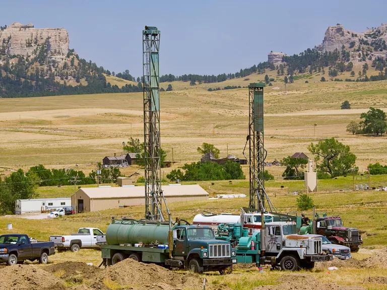Two tall rigs and other heavy equipment sit on a large expanse of land with mountains in the background