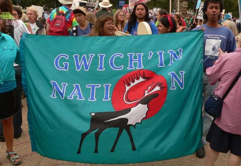 A woman at a protest holds a flag that reads "Gwich'in Nation"
