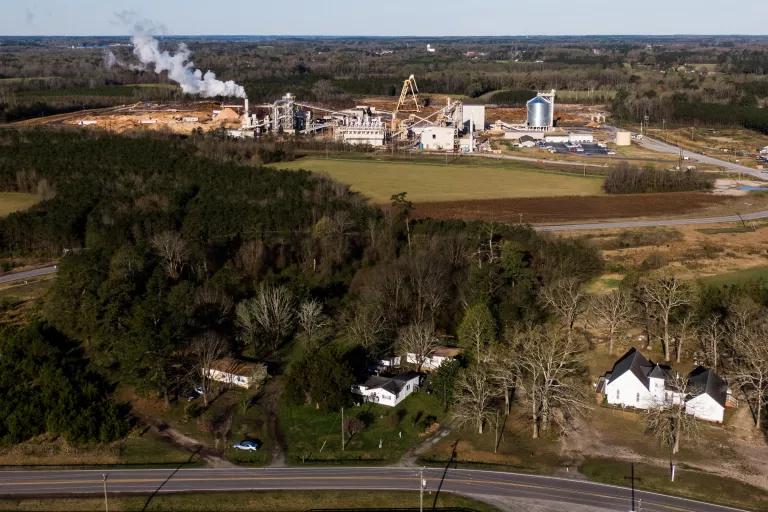 An aerial view shows smoke exiting a stack from a large industrial facility situated in a wooded area near residential homes