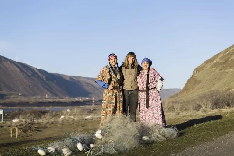 Three women stand in a valley with a river behind them