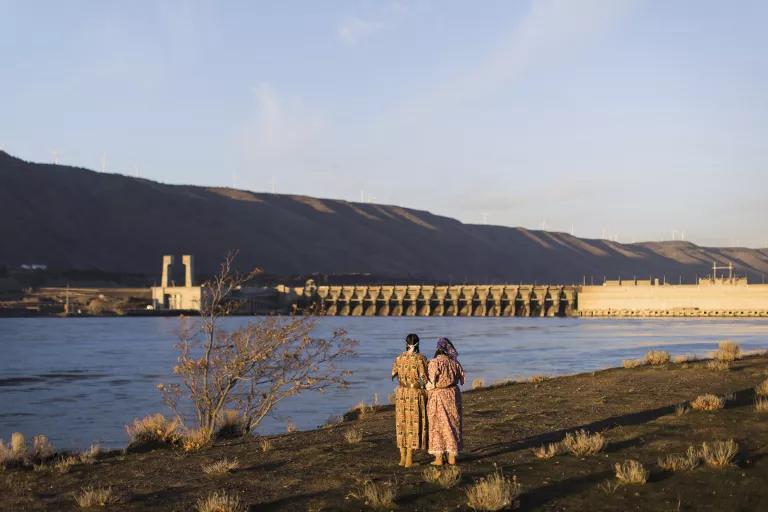 Two women stand at the edge of a river with a dam visible in the distance