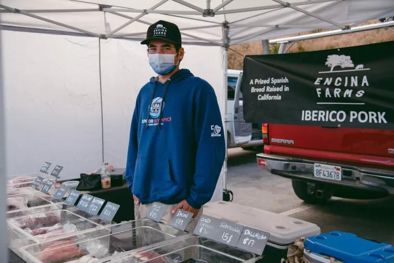 A man wearing a face mask stands behind a table displaying raw meat, with a pick-up truck behind him displaying a poster that reads "Encina Farms Iberico Pork"
