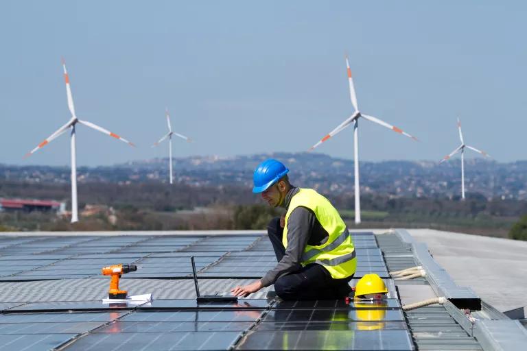 A worker sits on a large flat room among an array of solar panels, with wind turbines in the background