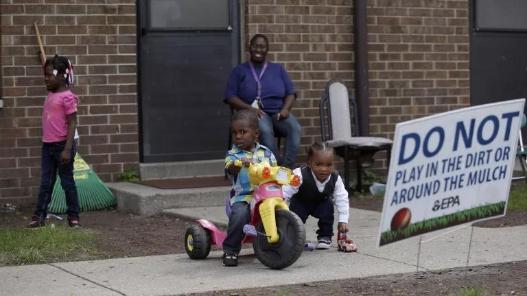 A mother sits outside of her apartment door as her two young sons play, one on a tricycle and the other with a toy car. The mother’s young daughter walks away past a rake leaning against the building. A sign in the dirt reads, “Do not play in the dirt or around the mulch. —EPA.”