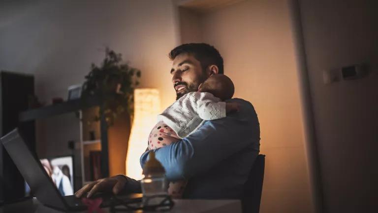 A man holds a sleeping infant in a dimly-lit room while working on a laptop