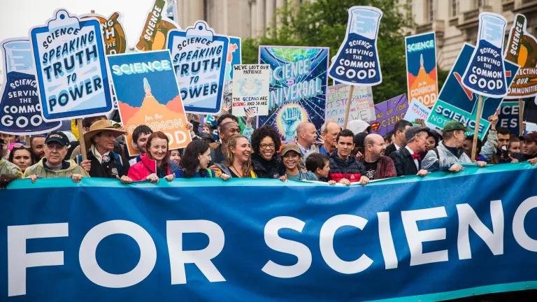 People march at a protest behind a large banner and holding signs that read "Science speaking truth to power" and "Science serving the common good"