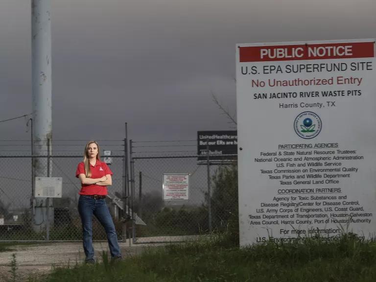 Alt Text: Jackie Medcalf stands next to a large U.S. Environmental Protection Agency Superfund site public notice sign for San Jacinto River Waste Pits in Harris County, Texas.