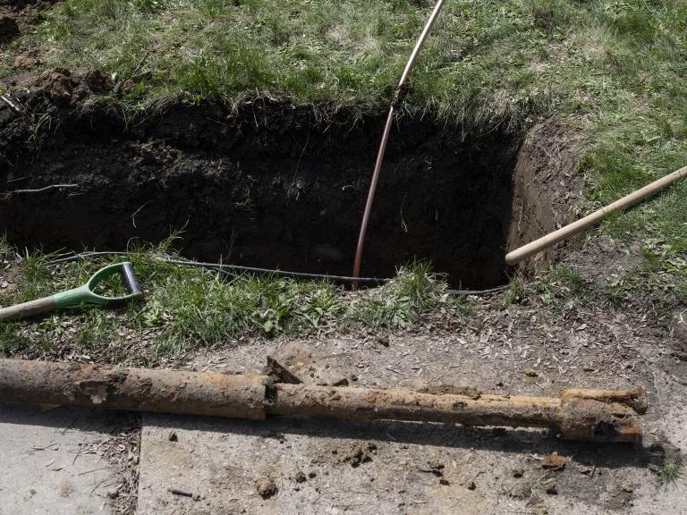 A new thin copper service line leads into a hole dug into the grass. Shovels and an old galvanized lead pipe lie around the hole.