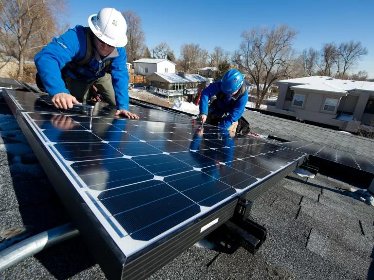 Two workers in hardhats install a solar panel on a house roof