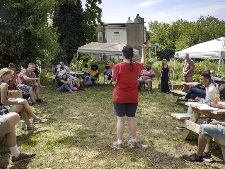A woman speaks to a group of people sitting outdoors at wooden tables 