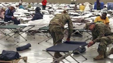 Two people in army fatigues set up rows of cots in a large space.