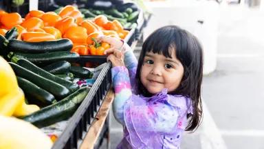 A young Latine girl reaching for an orange pepper on a stall at a Farmers Market in California.