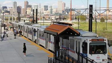 A light rail train pulls into a station with the Denver skyline seen in the background