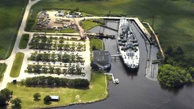 An aerial view of a battleship docked in a small inlet next to a tree-lined parking lot and building