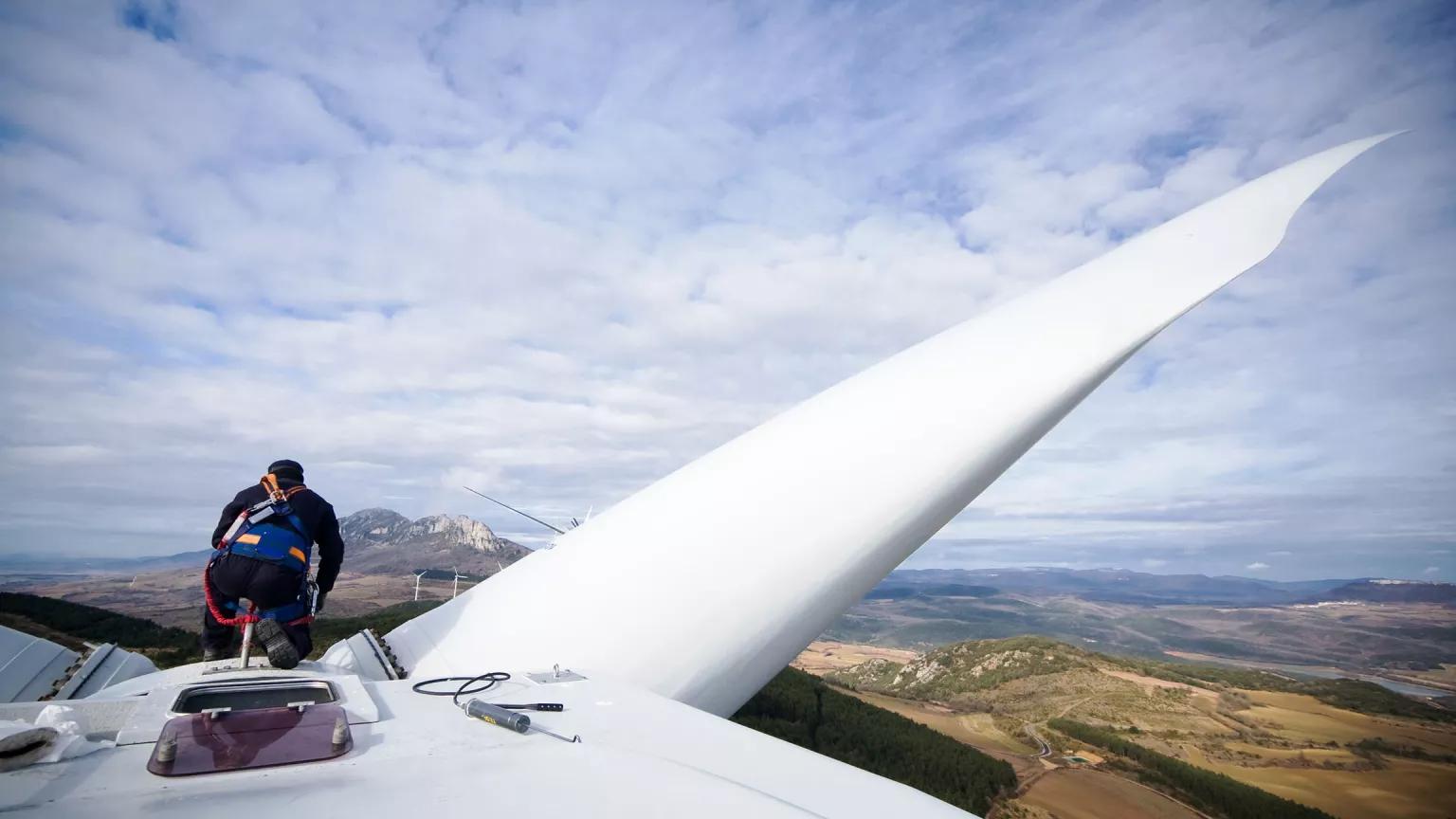 Rear view of a technician kneeling on the nacelle of a wind turbine and looking out at a sweeping, hilly landscape