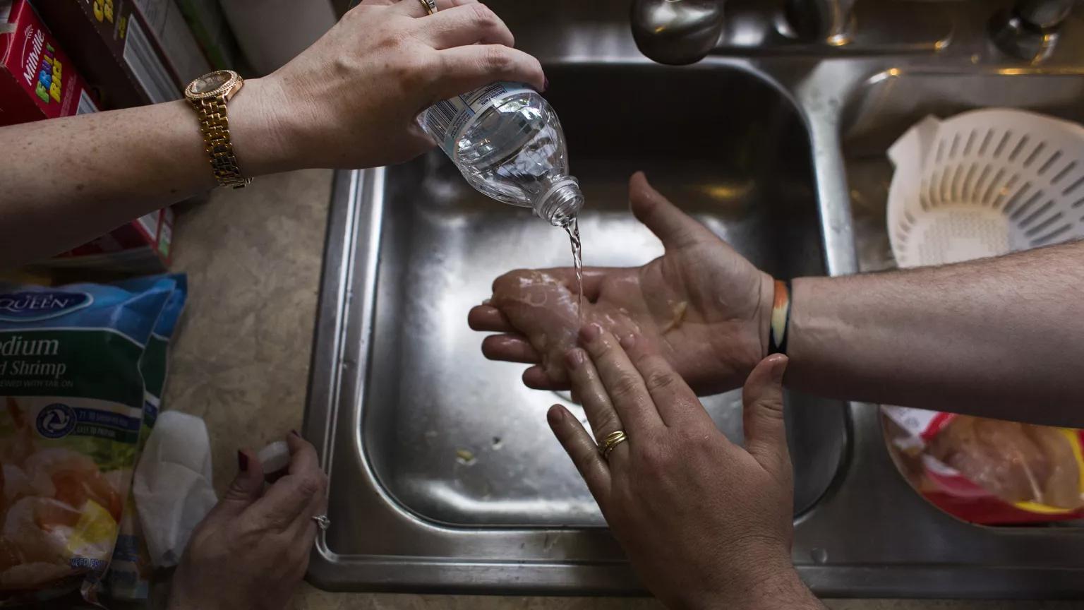 A person holds a piece of raw poultry over a kitchen sink, while another person pours bottled water over it.