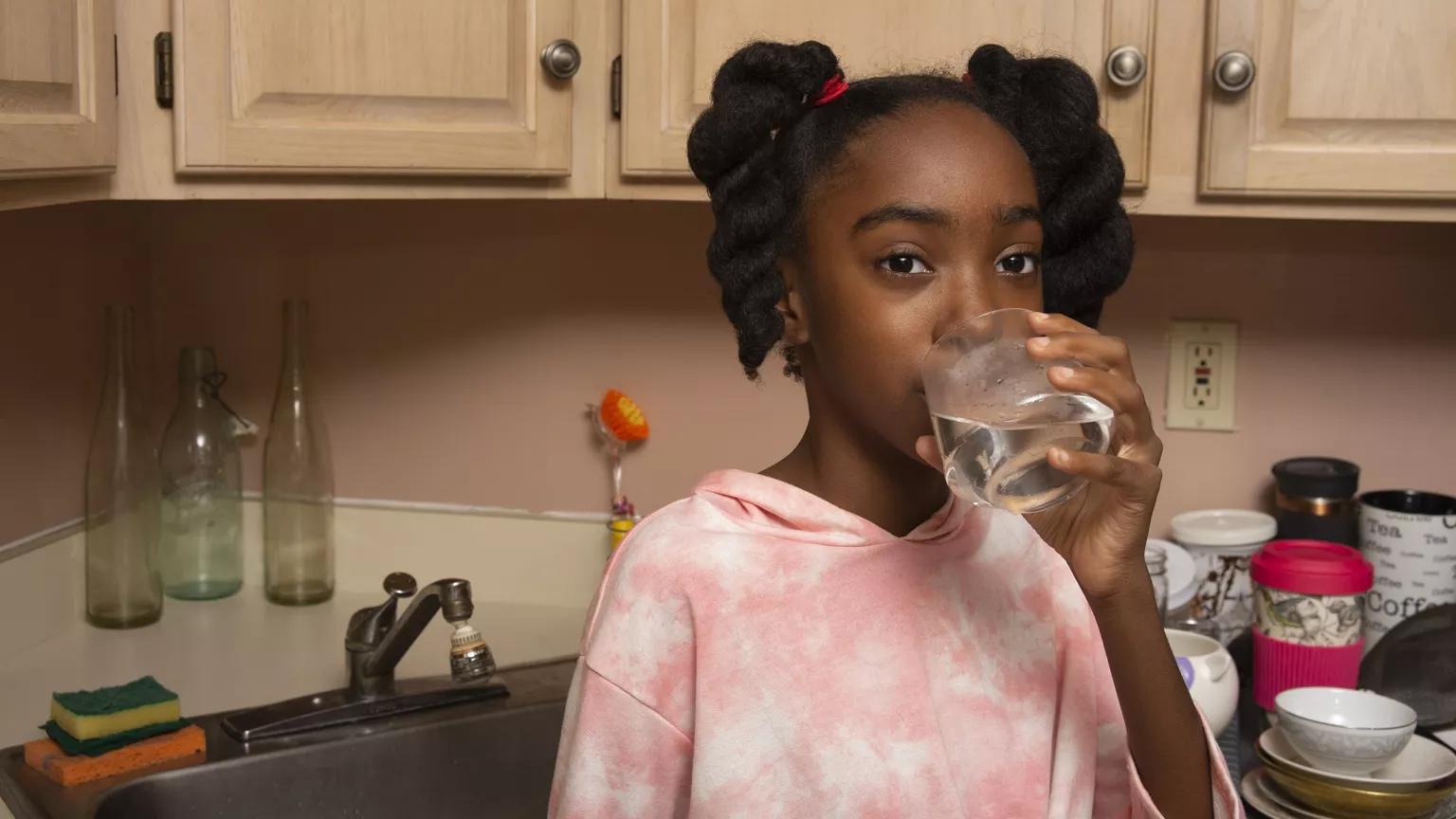 A Black pre-teen girl with braided pigtails and tie-dyed shirt is drinking a glass of tap water in front of a kitchen sink