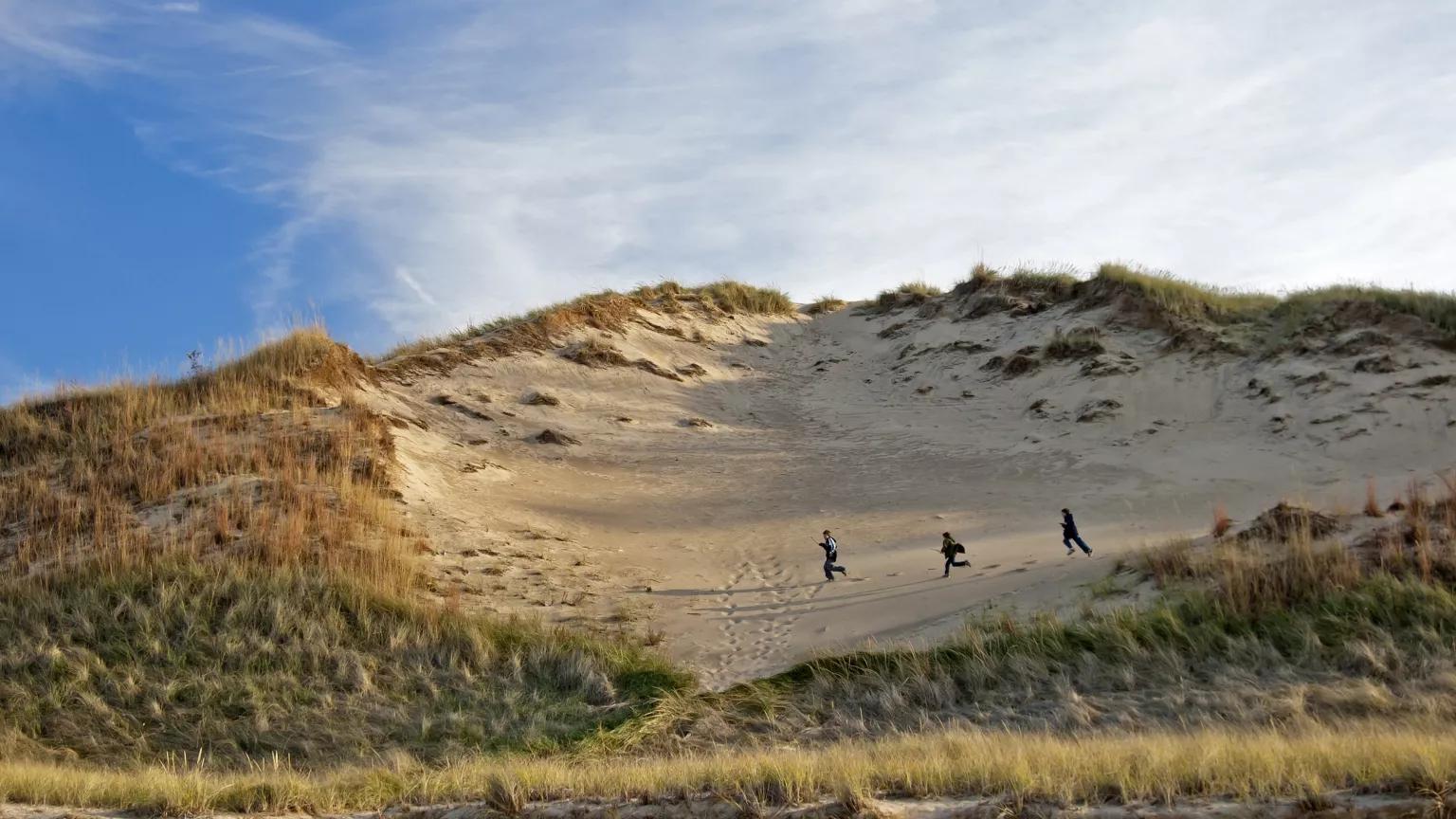 Three children run across a high sand dune with tall grasses