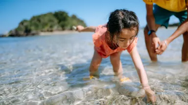 A child wading in clear water water picking up something from the water and holding an object in their other hand, with the legs of an adult behind her