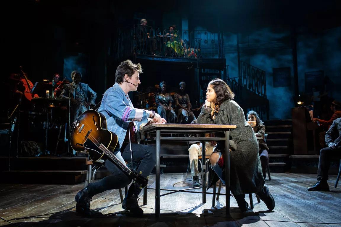 A man with a guitar and a woman sit at a table on a stage