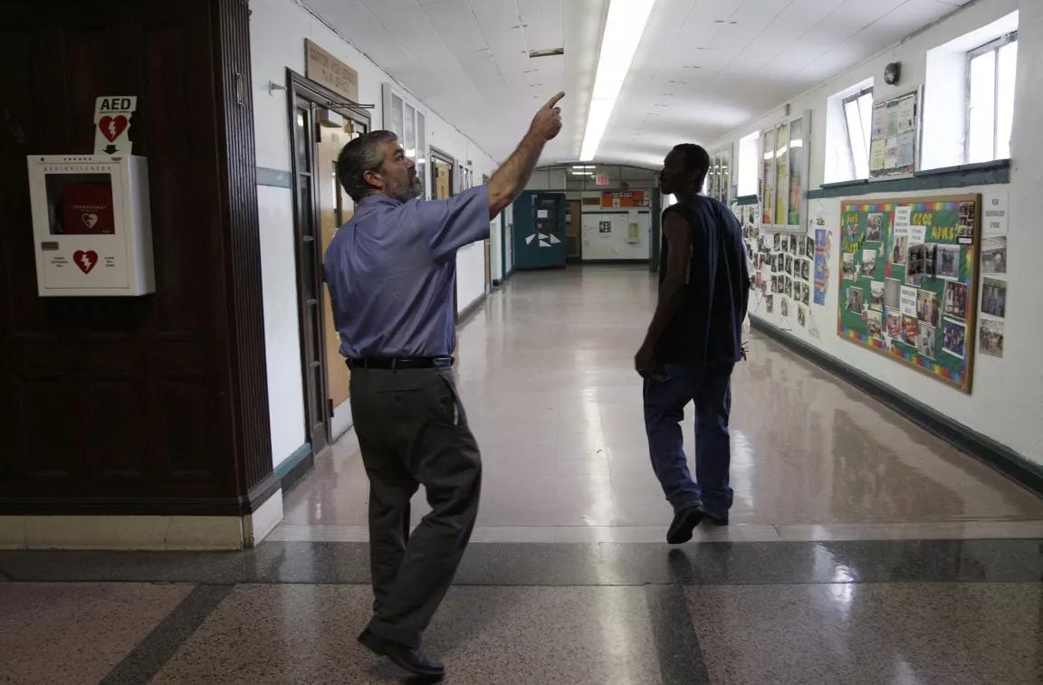 A person in a blue short-sleeve button down shirt and dark pants pointing at the ceiling of a school while another person walks slightly ahead of them