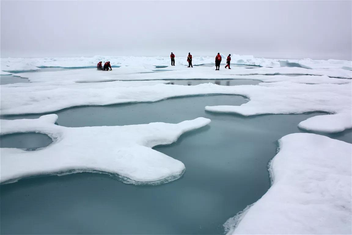 A group of people stand on a sheet of ice dotted with areas of water