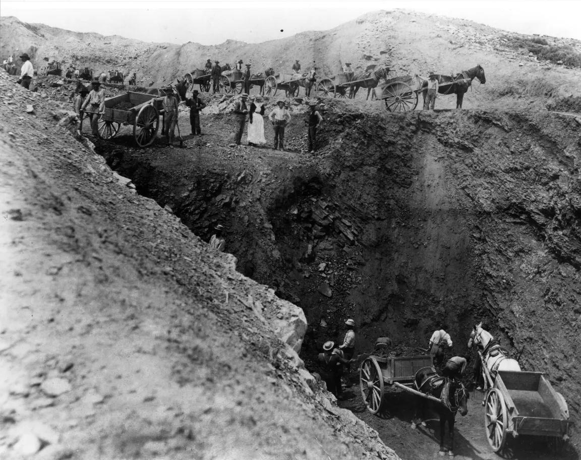 A black and white image shows people and horse-pulled carts surrounding a deep trench