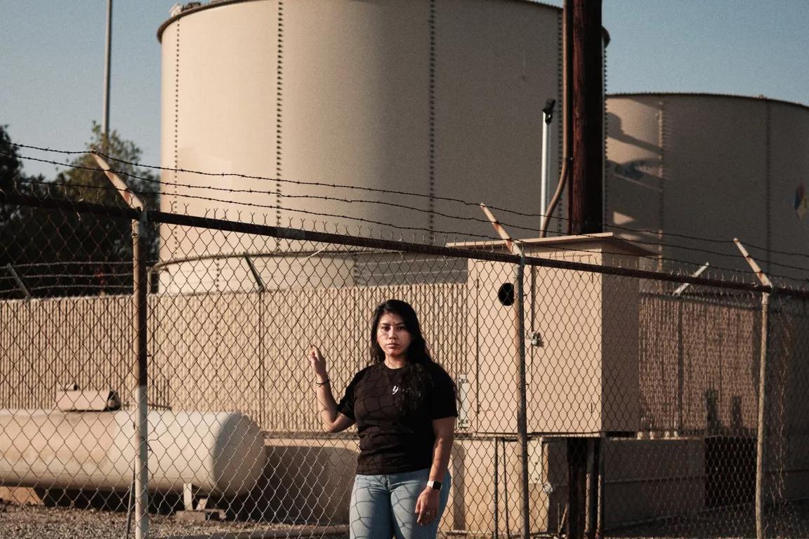 A woman stands in front of a chainlink fence topped with barbed wire, with large holding tanks on the other side