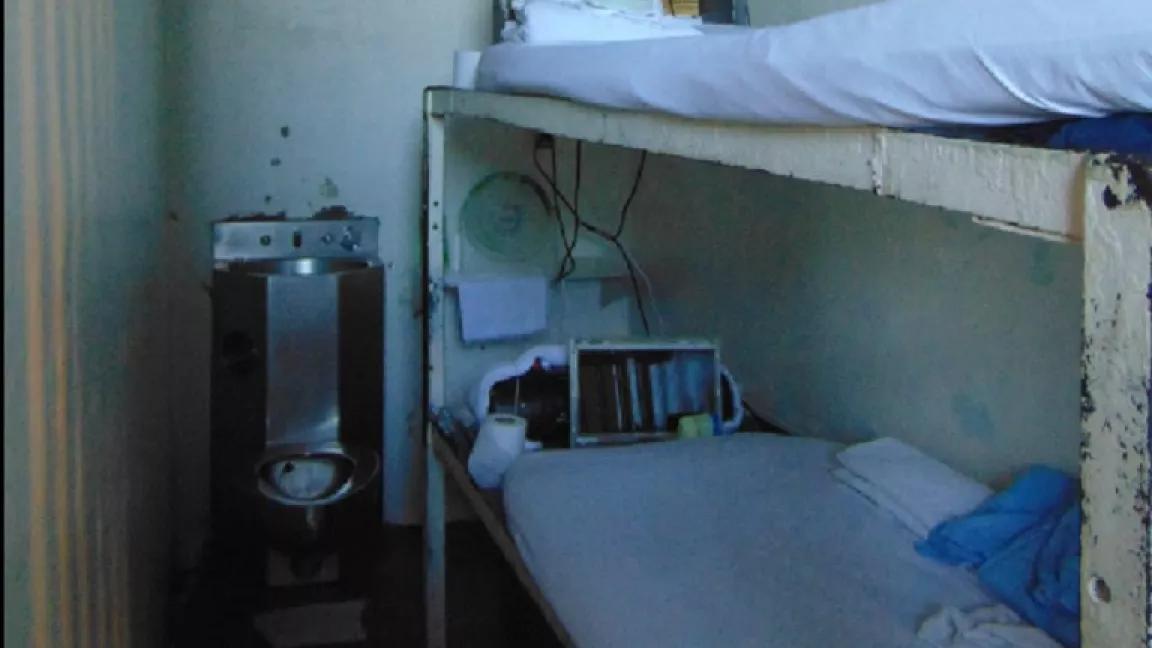 A prison cell containing a narrow bunk bed with an open toilet immediately next to it