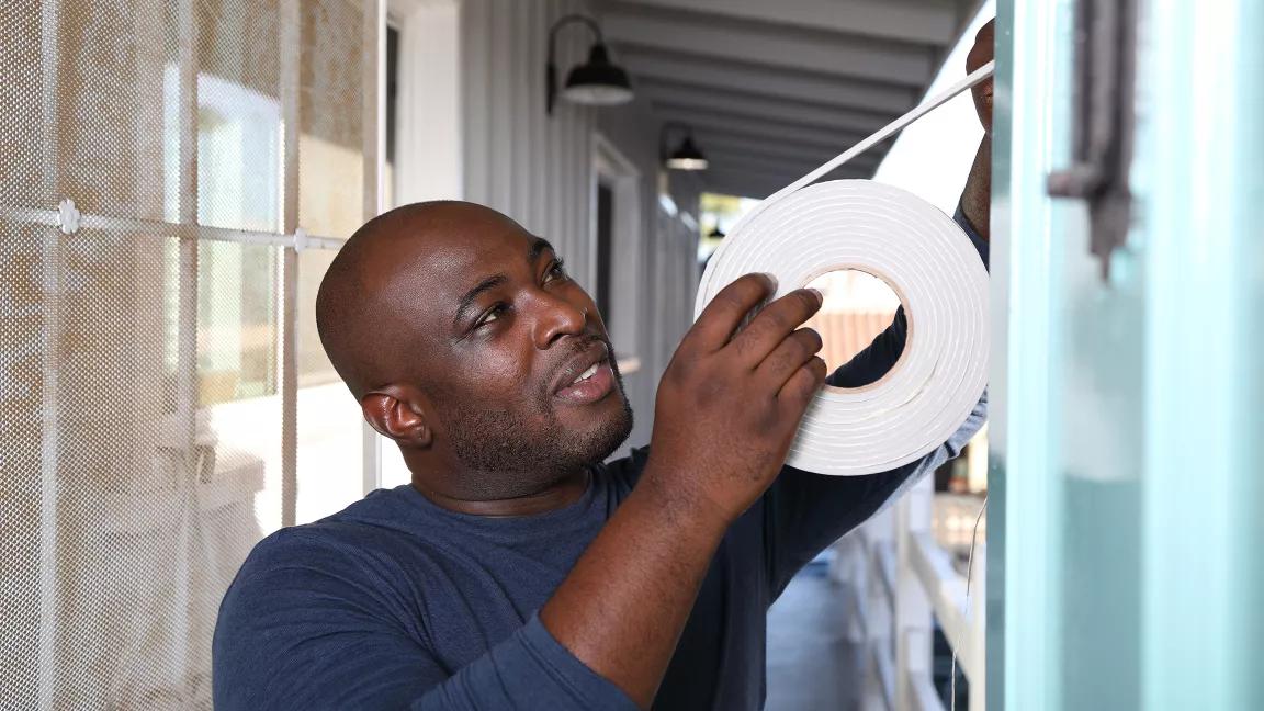 A Black man is applying a role of weather tape to an exterior door of his home. 
