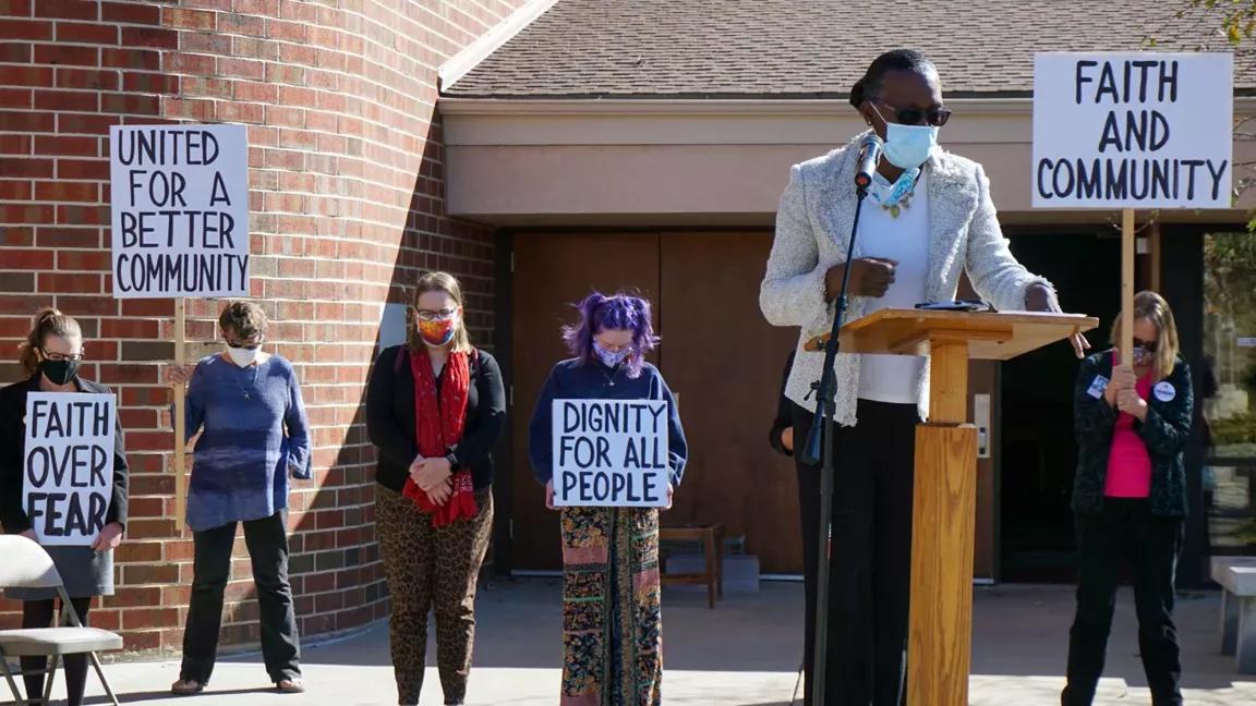 A woman speaks at a lectern in front of a brick building with people holding posters behind her that read "faith and community" and "united for a better community."