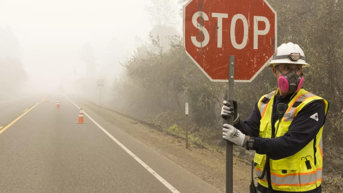 Photo of a person in a safety vest, hard hat, and respirator holding a stop sign in front of a road almost hidden by wildfire smoke. 