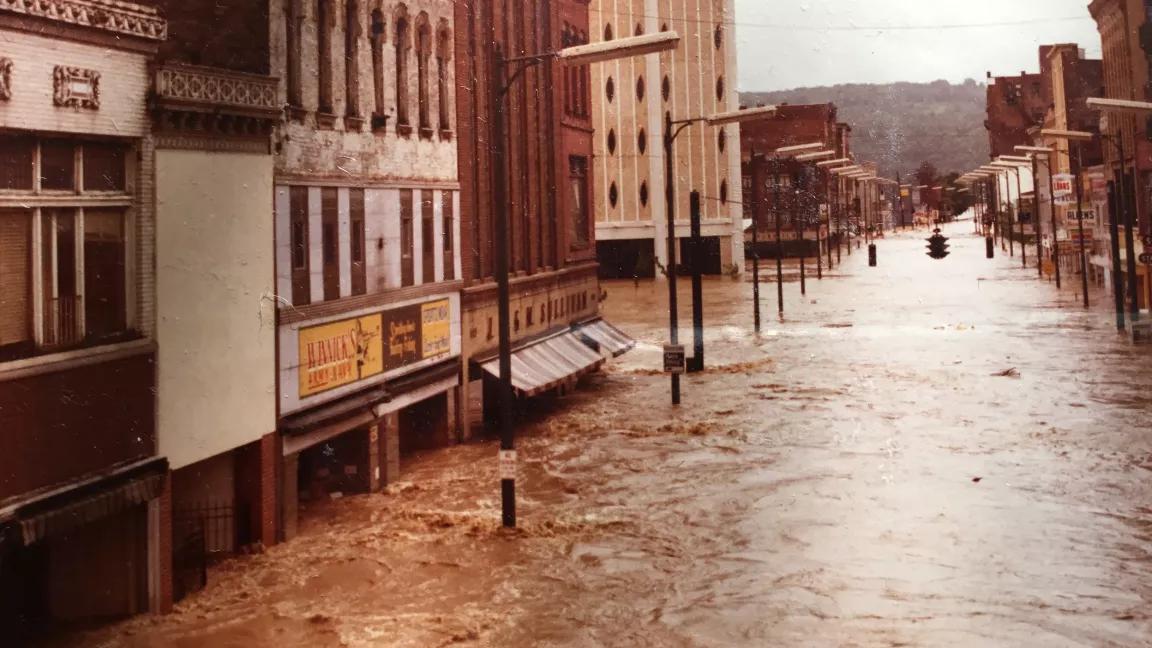 A 1970s-era photo shows a wide downtown city street with swirling brown floodwaters reaching most of the way up the first story of the buildings. 