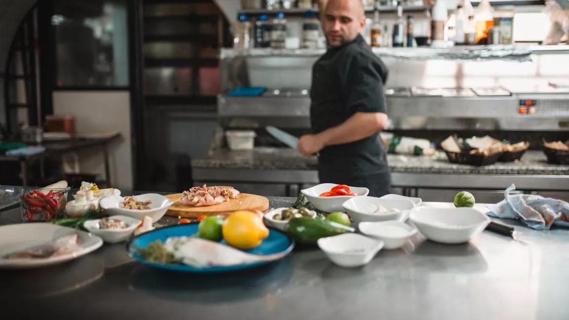 Chef in a black apron looks over a counter of freshly prepared food
