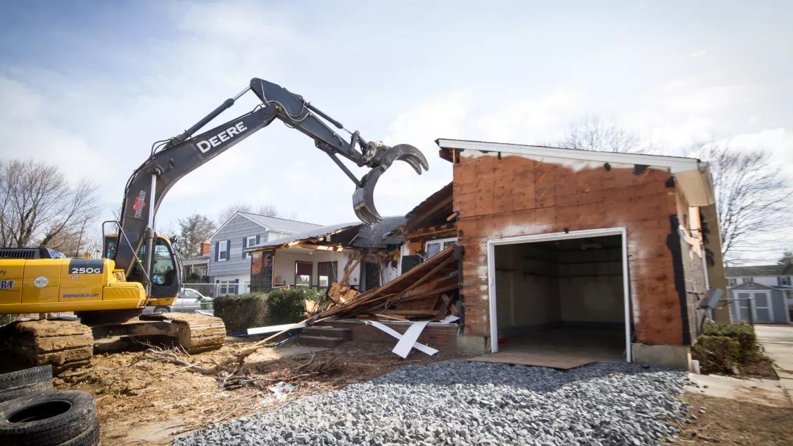 An excavator is in the process of demolishing a single-family home. A gravel driveway leads to a partially demolished garage, which is the only part of the home left standing.