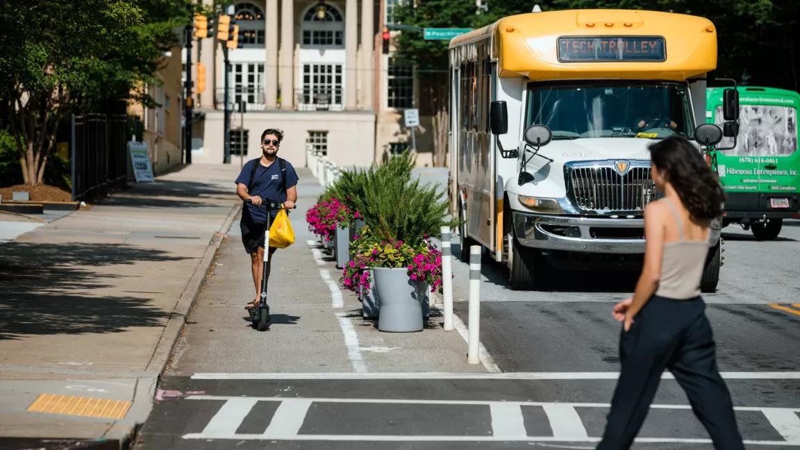A man rides a scooter in a protected bike lane in Atlanta, while a woman crosses the street and a University shuttle approaches 