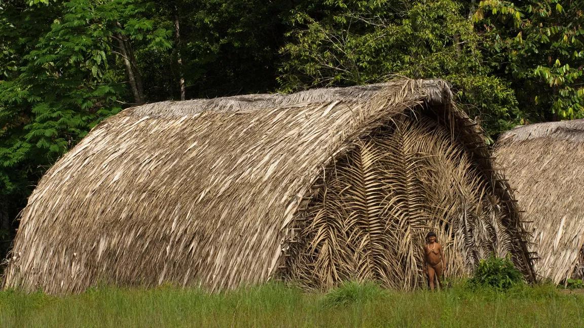 A woman stands in front of a large shelter made from thatched palm leaves