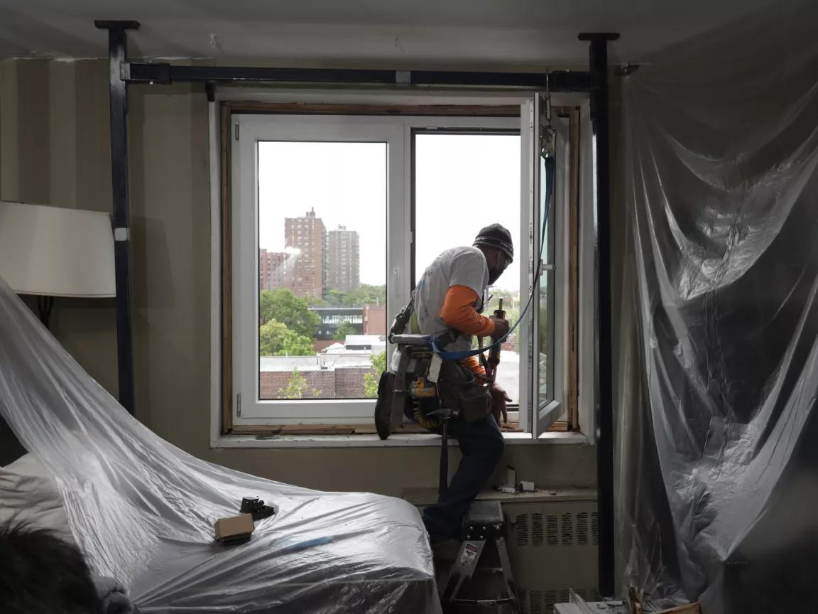 A man sitting on an open window of an apartment in the South Bronx, New York, preparing to install an energy-efficient window
