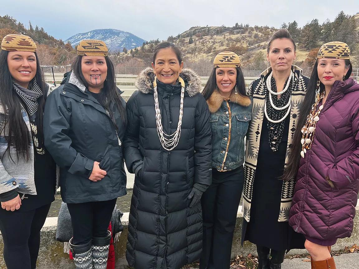 Six women stand in a row on the side of a paved road with mountains in the background
