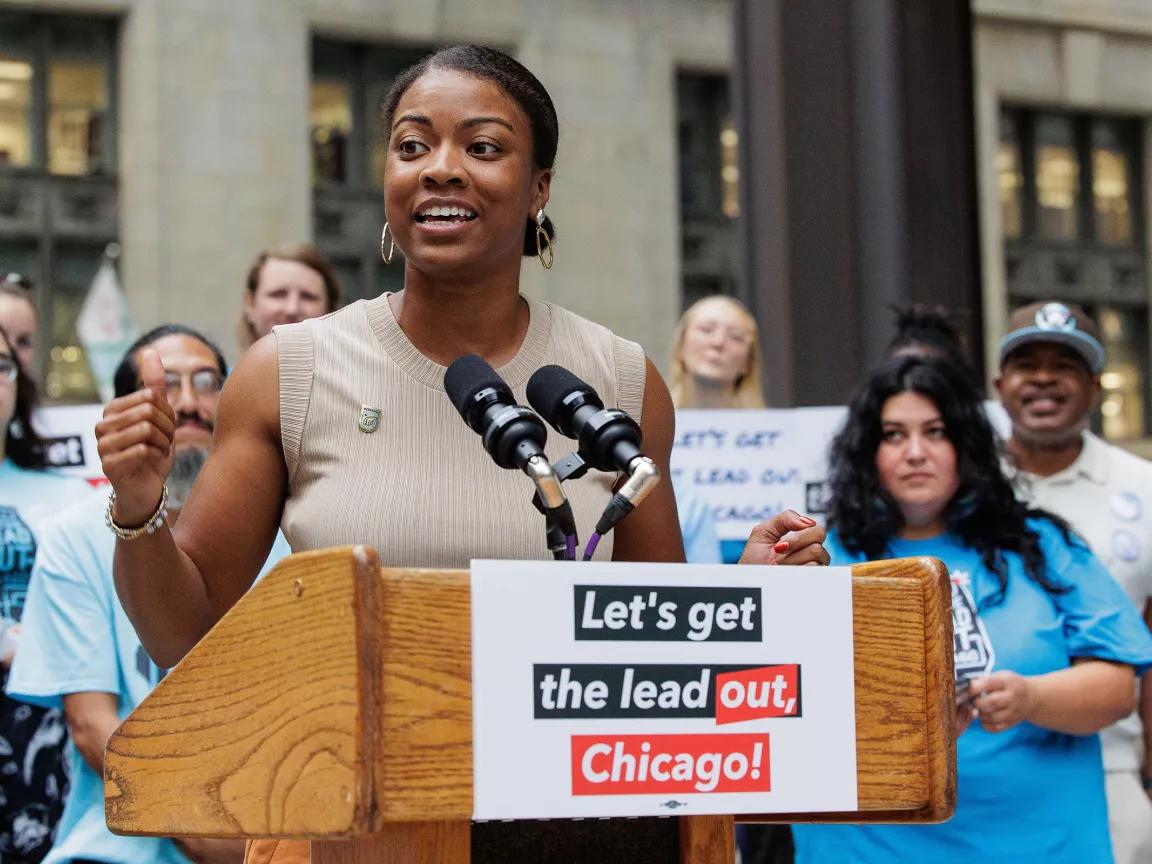 A woman speaking at a podium that has a sign that reads “Let’s get the lead out, Chicago!” with people standing behind her