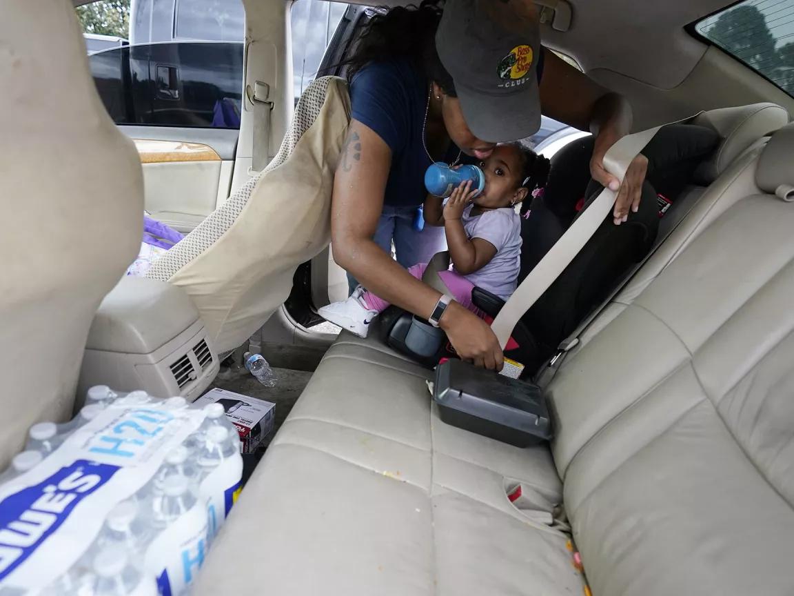 A Black woman leans over her daughter, who is drinking from a baby bottle, to buckle her seat belt. In the back seat of the car with the child is a case of bottled water.