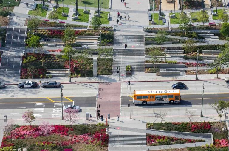A bus seen from above drives past on a street between two park blocks with zig-zagging paths amidst green space
