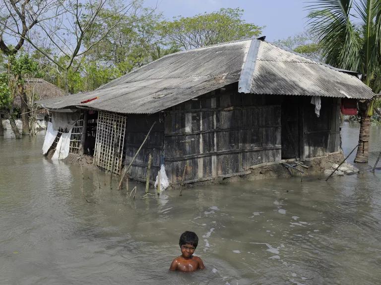 A child stands in deep floodwaters that are rising to the doorframe of a small house in the background