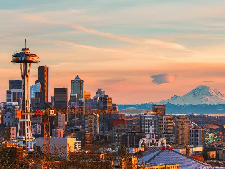 The sun sets over the Seattle skyline, including the Seattle Space Needle building and a snow-capped Mount Rainier