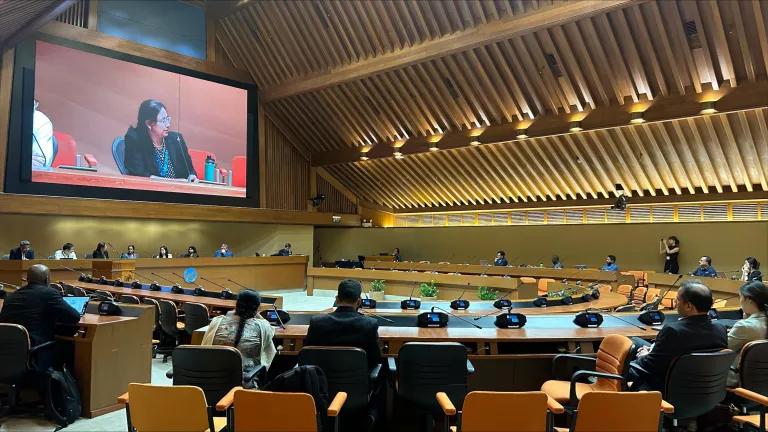 A large conference room with people seated along round tables with a giant screen showing the speaker