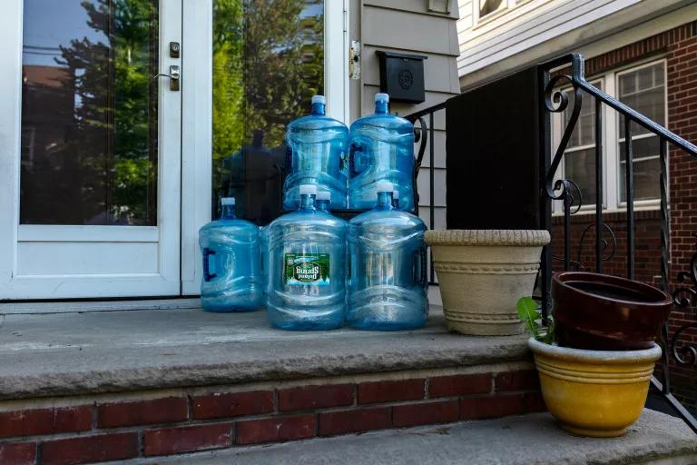 Large blue water jugs stacked near empty flower pots on the front porch of a house