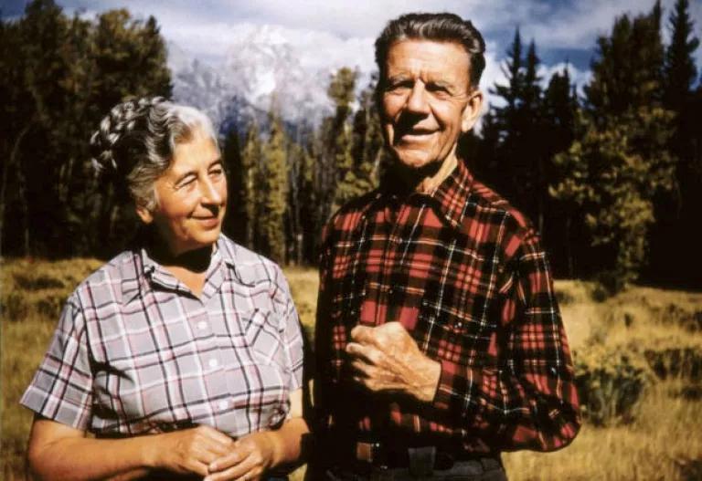 A man and woman stand in a clearing with trees and mountains behind them