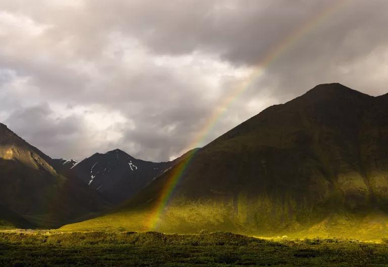 A rainbow rises from a grassy valley over mountains in the distance