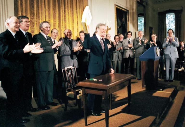 President Jimmy Carter stands near a small table holding a document high in the air, while people stand applauding behind him 