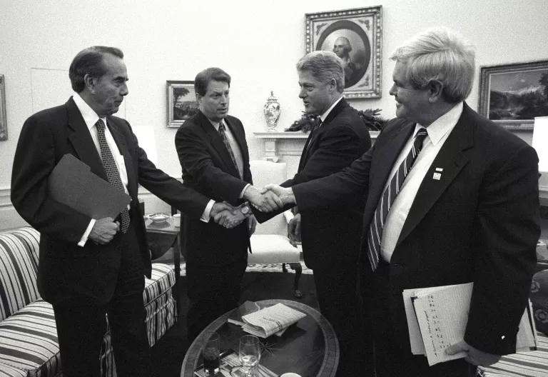 Senator Bob Dole, Vice President Al Gore, President Bill Clinton, and House Speaker Newt Gingrich stand in front of couches in the Oval Office and shake hands